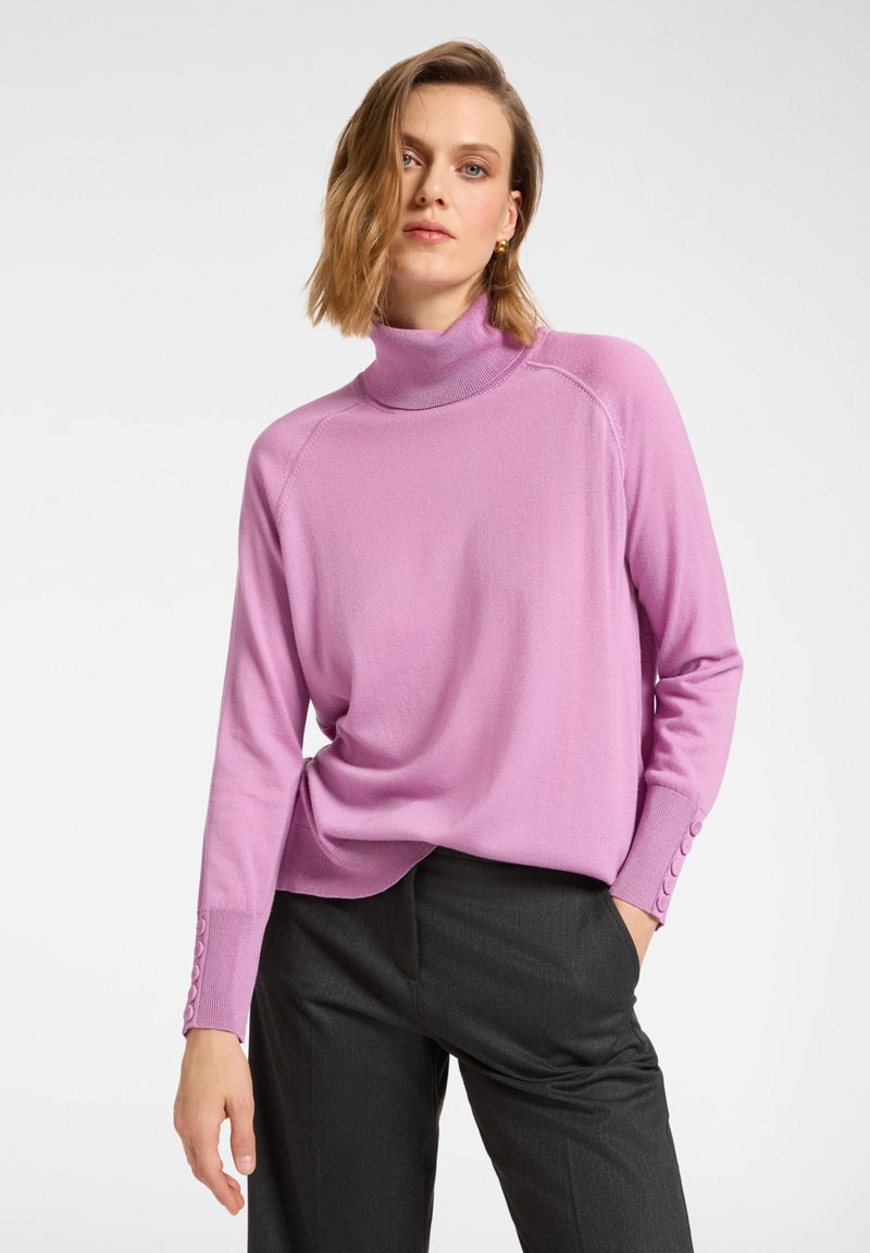 Różowy sweter z golfem i długimi rękawami, ozdobiony guzikami na mankietach, o luźnym kroju. Noszony z ciemnoszarymi spodniami.