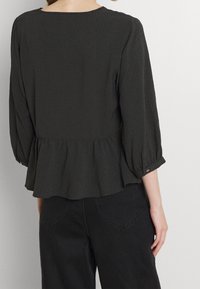 Top peplum grigio scuro in tessuto testurizzato, maniche a tre quarti e scollatura rotonda. Il retro presenta dettagli arricciati.