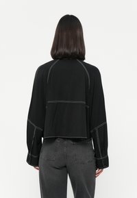 Black cropped blouse with long sleeves, featuring contrasting white stitching and a relaxed fit. Worn with dark gray high-waisted jeans.