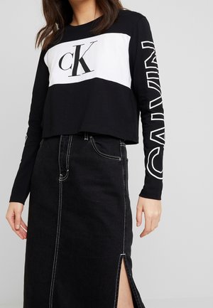 Mujer con top corto de manga larga negro con el logo blanco "ck" y texto "CALVIN" en la manga, combinado con falda negra de cintura alta con abertura frontal.