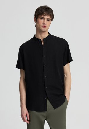 Giovane uomo che indossa una camicia nera a maniche corte con bottoni e pantaloni verde oliva, in piedi con una mano in tasca davanti a uno sfondo neutro.