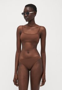 Brown ribbed bikini top with thin straps, paired with matching high-waisted bottoms. Model wears round black sunglasses against a plain backdrop.