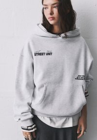 Haut à capuche gris avec une poche avant, présentant des motifs textuels en noir. Coupe décontractée, tissu doux, avec des rayures blanches et noires superposées au niveau des poignets.