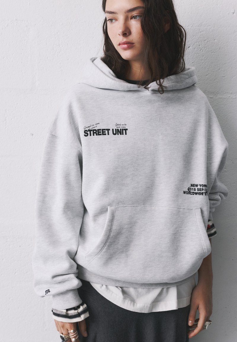 Haut à capuche gris avec une poche avant, présentant des motifs textuels en noir. Coupe décontractée, tissu doux, avec des rayures blanches et noires superposées au niveau des poignets.