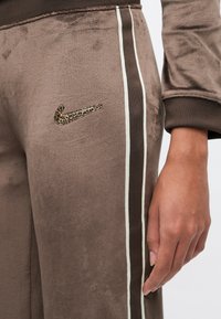 Καφέ παντελόνι από βελουτέ με λευκές και σκούρες καφέ ρίγες στο πλάι, λογότυπο Nike με λεοπάρ μοτίβο και ένα χέρι ακουμπώντας δίπλα στο πόδι.