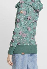 Felpa con cappuccio caratterizzata da un motivo floreale in verde acqua e viola, tessuto testurizzato, orlo verde a costine e una piccola etichetta in cotone marrone in basso.