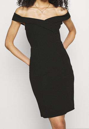 Femme portant une robe noire ajustée, épaules dénudées, les mains dans les poches, avec un collier en or orné d’un pendentif en forme de barre.
