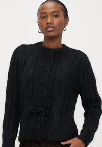 Μαύρο πουλόβερ πλεκτό με στρογγυλή λαιμόκοψη, υφή με ανάγλυφο σχέδιο, κλείσιμο με πέντε κουμπιά και ζώνη στη μέση. Υλικό με μείγμα μαλλιού.