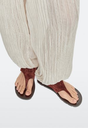 Füße tragen dunkelrote Wildleder-Zehenschlaufen-Sandalen, kombiniert mit hellen, plissierten, locker sitzenden Hosen vor schlichtem Hintergrund.
