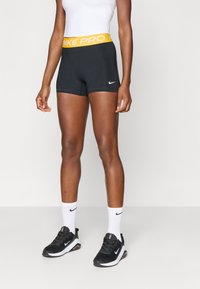 Fekete sportnadrág sárga Nike Pro derékpánttal, fehér Nike logóval. Fekete sportcipővel és fehér zoknival párosítva.