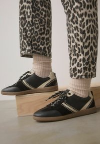 Schwarze und beige Sneaker mit einer strukturierten Oberseite, kontrastierenden Nähten und gemusterten Schnürsenkeln, kombiniert mit Jeans im Leopardenmuster und gerippten Socken.