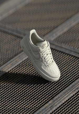 White Nike Air Force 1 sneaker featuring smooth leather, perforated toe, tonal laces, and logo accents in light gold. Floating above a textured surface.