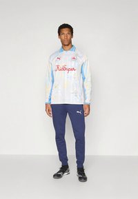 Világoskék hosszú ujjú mez virágmintával és piros "KidSuper" felirattal; sötétkék melegítőnadrággal és fekete sportcipővel párosítva.