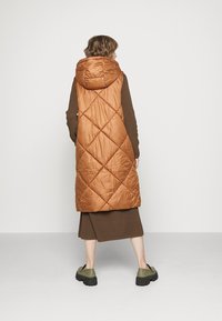 Pitkä, hihaton, tikattu liivi lämpimän ruskeassa värissä, jossa on huppu. Siinä on timantinmuotoiset tikit, ja se on päällä ruskean neulotun asun päällä.