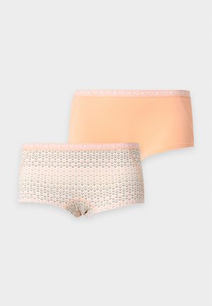 Γυναίκα στέκεται ξυπόλητη, φοράει λευκό crop top και βερμούδα σε ροδακινί χρώμα με απλό φωτεινό φόντο.