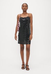 Nő fekete, flitteres mini ruhában, vékony pántokkal, fekete magassarkú szandálban, és egy rózsaszín flitteres válltáskával.