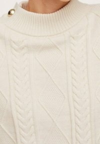 Pull crème en maille torsadée avec un col côtelé, présentant des motifs diagonaux et tressés, et un bouton décoratif doré près de l'encolure.
