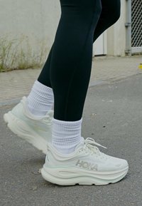 Pessoa vestindo leggings pretas, meias brancas de tecido canelado e sapatilhas de corrida HOKA brancas, em pé sobre uma superfície pavimentada ao ar livre.