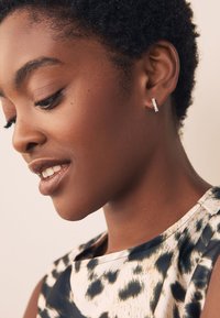 Silberner Creolenohrring mit rechteckigem Design und klaren Steinakzenten, getragen von einer Person mit kurzem, strukturiertem Haar und einem gemusterten Oberteil.