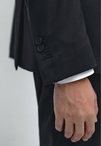 Ærme på mørk jakkesætjakke med dobbelte sorte knapper og syningsdetalje. Hvid skjortemanchet synlig under. Hånd hvilende ved siden.