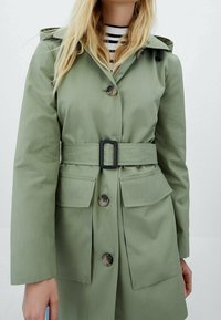 Light green waterproof jacket with a belt, button closure, large pockets, and a hood. Smooth texture and relaxed fit.