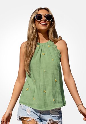 Lachende vrouw met oversized zonnebril, groene mouwloze top met gele bloemrijke borduursels en versleten lichte denimshorts.