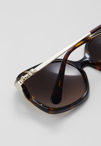 Occhiali da sole tartarugati con lenti scure sfumate, dettagli in metallo dorato e un'accessorio decorativo sulla tempia.