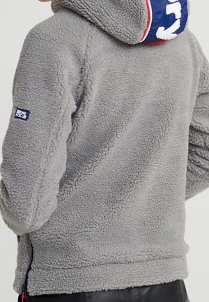 Person trägt eine graue Fleecejacke mit Kapuze, die ein blau-rotes Design und ein kleines rechteckiges Logo am Ärmel aufweist.