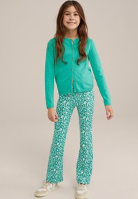 Tirkīza krāsa cardigan ar zeltītām pogām, apvienots ar zaļiem platiem biksēm ar baltiem ziedu rakstiem. Apģērbu papildina sporta apavi.