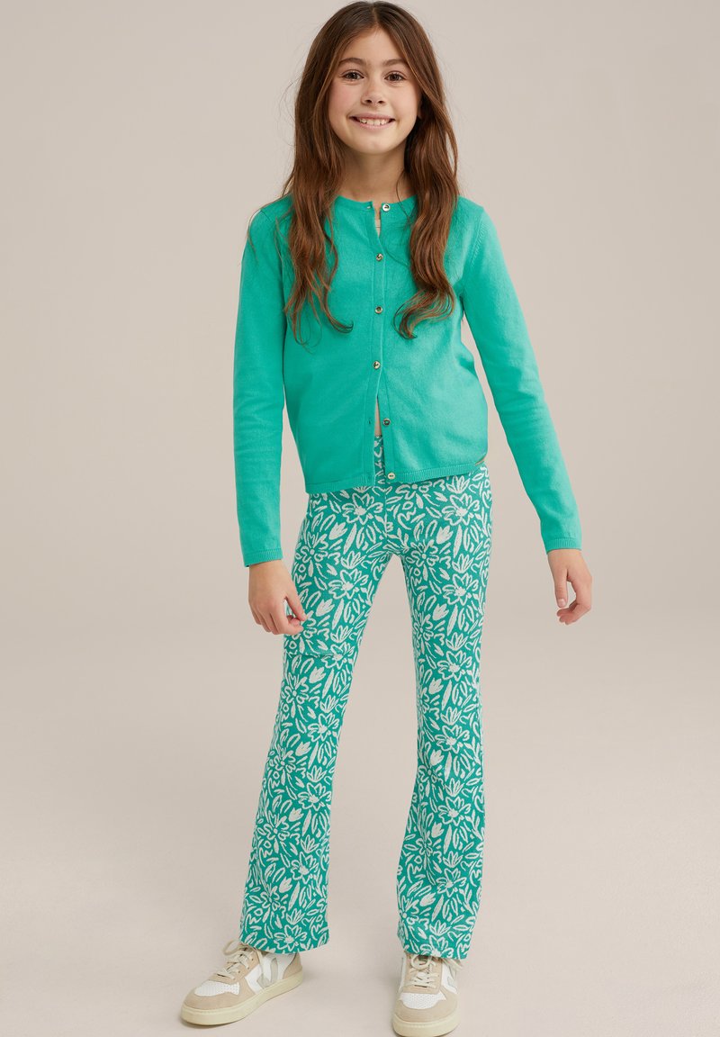 Tirkīza krāsa cardigan ar zeltītām pogām, apvienots ar zaļiem platiem biksēm ar baltiem ziedu rakstiem. Apģērbu papildina sporta apavi.