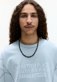 Světle modrý svetr s potiskem textu, doplněný dvěma náhrdelníky z korálků - jedním neprůhledným a jedním barevným. Viditelné přirozené kudrnaté vlasy.