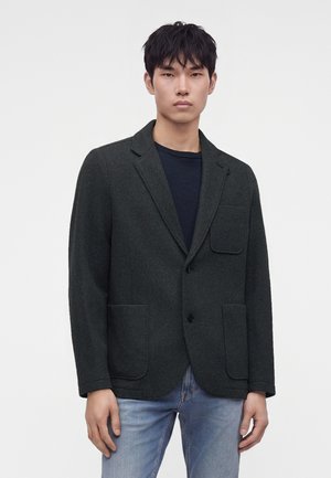 Jeune homme portant un blazer en laine vert foncé avec de grandes poches plaquées sur une chemise noire et un jean bleu clair sur fond blanc.
