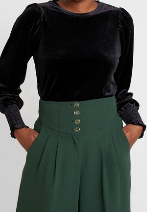 Persoon draagt een zwarte fluwelen top met lange mouwen en gerimpelde manchetten, gecombineerd met donker groene broek met hoge taille en vier gouden knopen. Handen in de zakken.