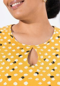 Geel shirt met ronde hals en een patroon van witte en zwarte stippen, met een sleutelgat uitsnede bij de halslijn en een textuurstof.