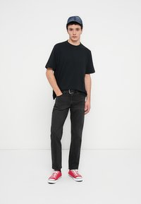 Μαύρο υπερμεγέθες t-shirt, γκρι τζιν και κόκκινα αθλητικά παπούτσια. Ο φορέας φοράει καπέλο από τζιν, με κοντά σγουρά μαλλιά και ουδέτερη έκφραση.