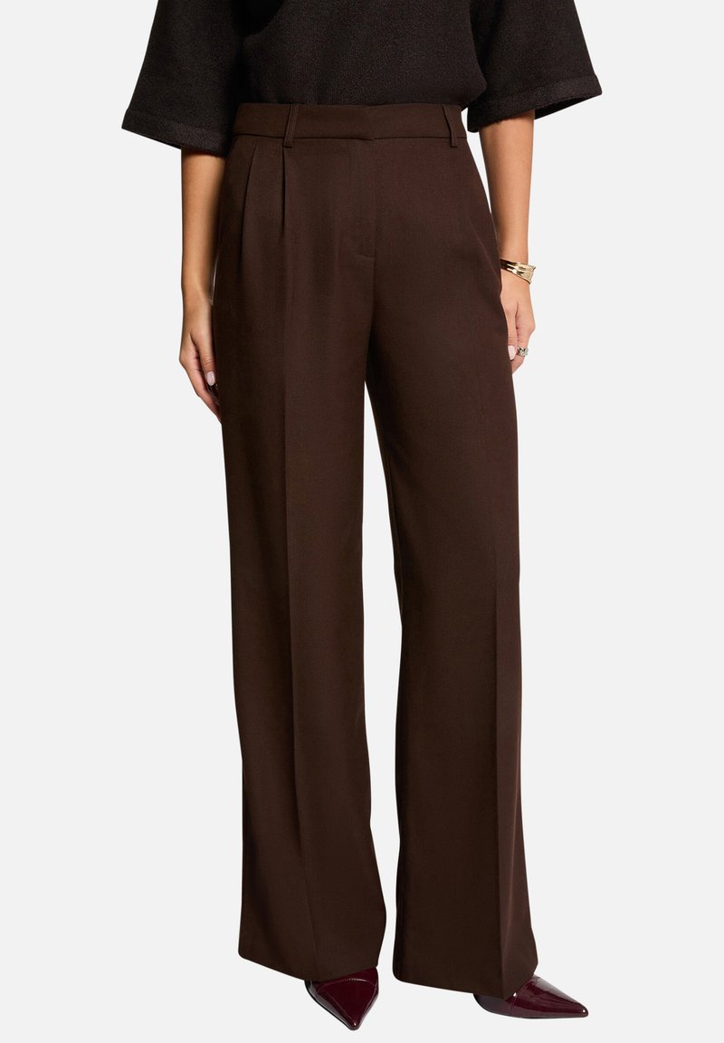 Pantalons larges marron au design ajusté, avec des plis sur le devant et une texture de tissu lisse. Portés avec des talons à bouts pointus.