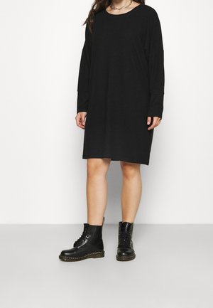 Kobieta ubraną w luźną czarną sukienkę z długim rękawem, czarne sznurowane botki za kostkę oraz srebrny łańcuszek na szyi na jednolitym białym tle.