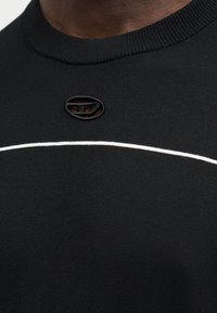 Κοντινή λήψη ενός μαύρου πλεκτού πουλόβερ με ένα μικρό οβάλ λογότυπο πάνω από μια οριζόντια λευκή ρίγα στο στήθος.