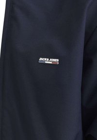 Giacca blu navy realizzata in tessuto liscio, con un piccolo logo argentato "JACK & JONES" e accenti rossi e blu sul lato sinistro del petto.