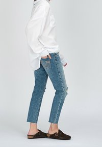 Biała koszula z długim rękawem, jasnoniebieskie dżinsy z przetarciami oraz brązowe zamszowe klapki. Dżinsy mają prosty krój i postrzępiony dół.