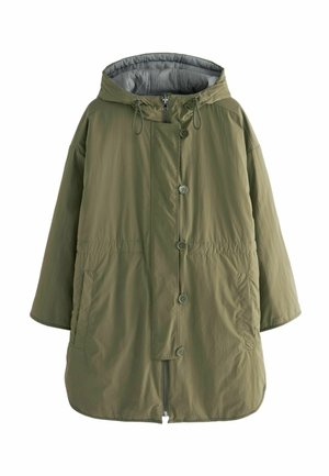 Personne portant un parka vert par-dessus une chemise rayée noir et blanc et un jean bleu, debout à l'extérieur dans une zone herbeuse.
