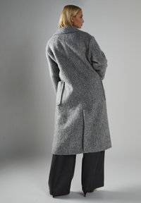 Grauer Woll-Langmantel mit strukturiertem Finish, großem Kragen und schrägen Seitentaschen. Über weiten schwarzen Hosen getragen.