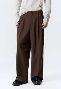 Pantalon marron à jambes larges avec une taille haute, des plis et une fermeture à un bouton, fabriqué dans un tissu lisse. Porté avec un haut léger à manches longues.