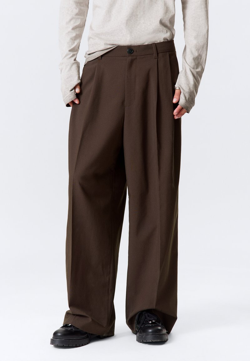 Pantalon marron à jambes larges avec une taille haute, des plis et une fermeture à un bouton, fabriqué dans un tissu lisse. Porté avec un haut léger à manches longues.