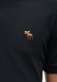 Μαύρο υφασμάτινο με υφή, με ένα μικρό κεντητό καφέ άλγος με κίτρινα κέρατα κοντά στην περιοχή του στήθους ενός πουκάμισου.