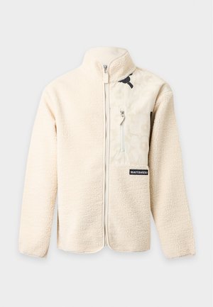 Homme portant une veste polaire crème, une chemise rayée, un pantalon noir à fleurs, des chaussures noires, portant un sac cabas rose clair à motifs sur l'épaule.