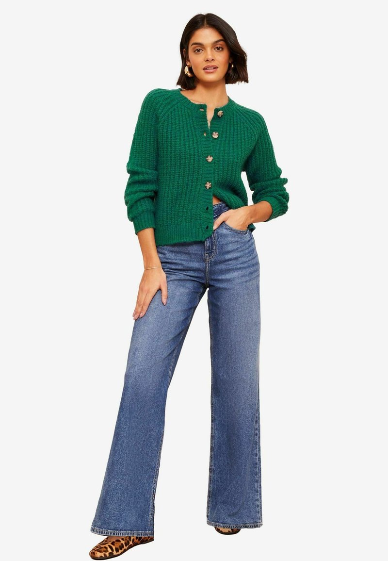 Grüne gerippte Cardigan mit Knopfverschluss, langen Ärmeln und rundem Halsausschnitt, kombiniert mit weit geschnittenen blauen Jeans und Schuhen im Tiermuster.