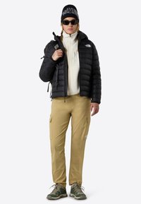 Svart pufferjacka över en ljusbeige fleece, khakifärgade cargo-byxor och grågröna vandringsskor. Tillbehör inkluderar en svart mössa och solglasögon.