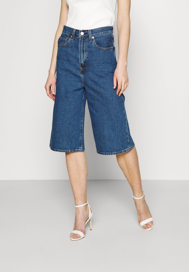 Femme portant un short en jean bleu jusqu'aux genoux, associé à des sandales blanches à talons hauts et un top blanc sans manches.