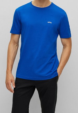 Man wearing a plain bright blue short-sleeve T-shirt with "BOSS" logo on chest, paired with black pants, standing with one hand in pocket.